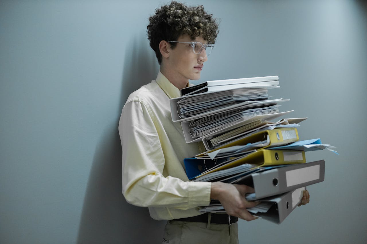 services-01 An overworked office worker carrying a large stack of files and folders in a modern setting.
